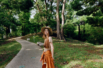 Fototapeta premium Woman walking in park on gravel path among tall trees, wearing a wide hat and orange skirt, smiling at camera. Outdoor nature portrait, summer leisure and relaxed lifestyle scene.