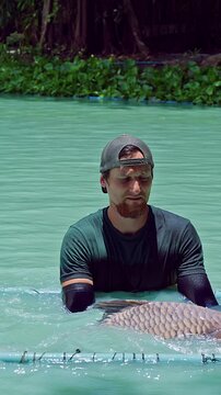 Vertical video. Fisherman Standing In Turquoise Lake Holding Large Siamese Carp Over Floating Frame With Tropical Plants And Fountain In Background, Catch And Release Fishing