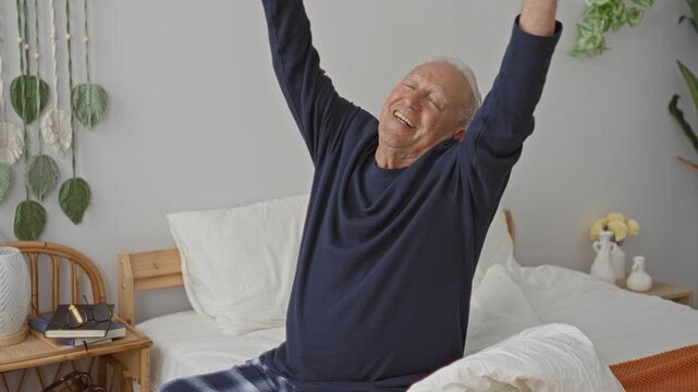Elderly man stretching arms while sitting on bed in building, wearing pajamas, smiling and reaching overhead toward ceiling; morning contentment.