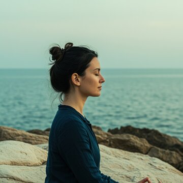 Serene woman meditating in profile on a rocky coastline with ocean background for mindfulness app marketing, wellness blogging and mental health advertising