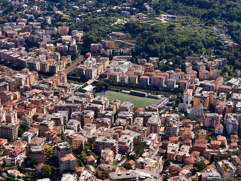 Aerial view of the urban landscape featuring dense residential buildings, a green sports field, and lush hillsides under bright daylight Rapallo, Liguria, Italy.