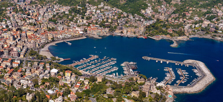Aerial view of the harbor and coastal town of Rapallo with colorful buildings, yachts moored in the marina, and lush green hills under bright sunlight in Rapallo, Liguria, Italy.