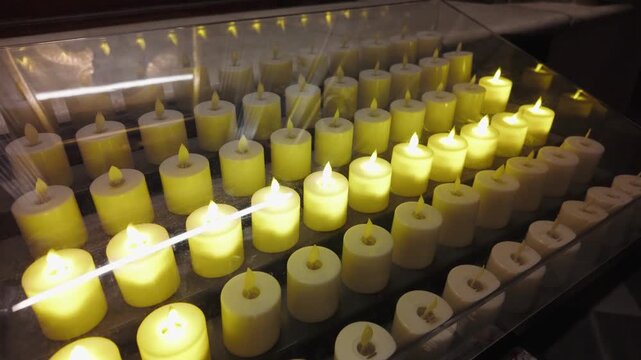 Electric votive candles glowing with warm yellow light arranged in neat rows inside a transparent glass display case within a peaceful church setting for prayer meditation and religious spiritual 