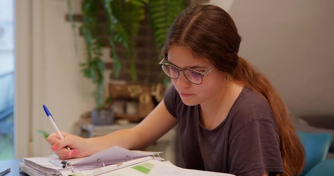 Focused Student Studying: A young student, engrossed in her studies, leans over a desk, diligently writing with a pen, surrounded by books and papers.