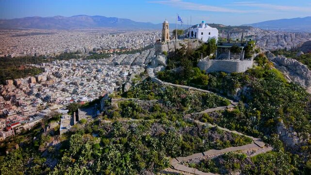 Aeriaof the Lycabettus Hill summit in Athens, showing the St. George Chapel, the peak viewpoint and the Greek flag overlooking the vast urban landscape under a clear midday sky.