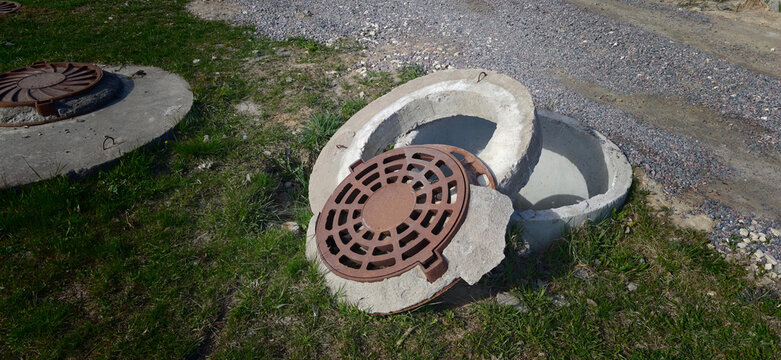 Broken concrete manhole cover lying beside an open sewer access point on a gravel road surrounded by green grass and dirt