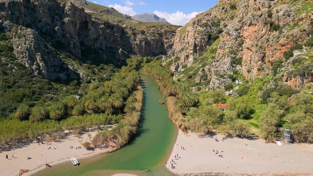 Aerial view of Preveli Beach and heart-shaped palm river in Crete, Greece. Drone fly back from palm forest canal to open sea, sandy beach and mountains. Scenic tropical landscape in Europe 4k