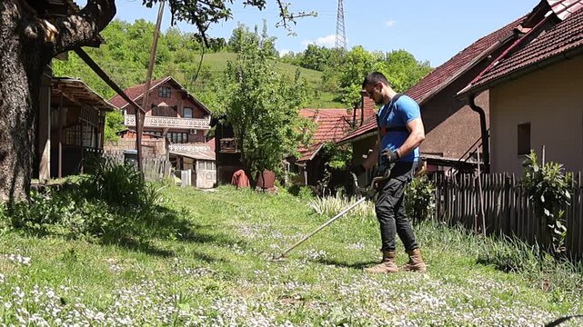 Focused worker trimming grass with a string trimmer in a village yard, maintaining neat edges along the fence line, with bright daylight and lush greenery emphasizing outdoor home care tasks.