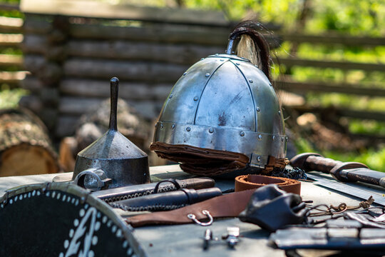 Historical steel helmet with horsehair crest and leather lining, placed on a table next to a metal shield boss, sword, and leather accessories, showcasing armorer craftsmanship in natural sunlight wit