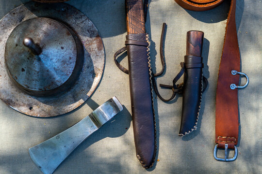 Top down view of medieval equipment: steel shield boss with rust, shiny axe blade, two knives in leather sheaths, and a brown belt with buckle, arranged on grey canvas in natural lighting with clear m