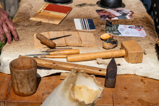 Collection of traditional craft tools on a fur-covered table, wooden mallets, metal hammer, chisels, and a wooden cup, background with bark pieces, shells with pigment, and flyers, natural daylight, d