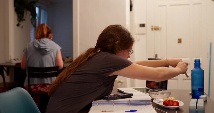 Daily life focus: A woman engages in daily activities, her attention is captured by objects on the table, while another figure is blurred in the background, set within a residential indoor setting.
