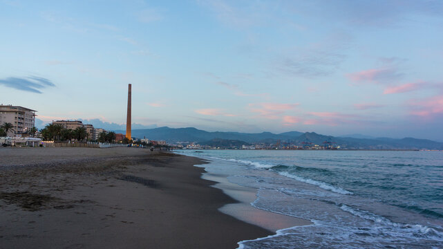 Evening view of Malaga beach with historic industrial chimney, calm sea and pastel sky.
