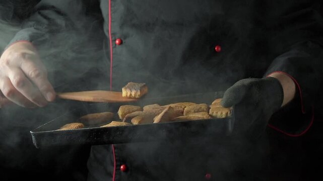 Chef in black uniform lifts tray of freshly baked cookies, using wooden spatula to serve them while steam rises in a cozy kitchen setting