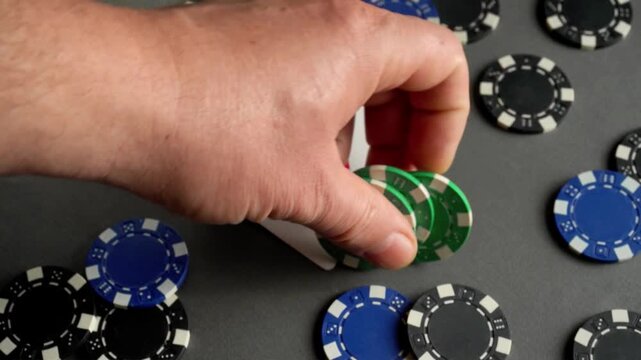 A hand reaches for green poker chips and takes them from an Ace playing card. The remaining chips are scattered across a gray surface, creating a dynamic gaming scene
