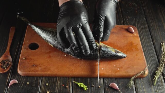 Chef in black gloves skillfully fillets mackerel fish on wooden cutting board with herbs and spices arranged around the scene