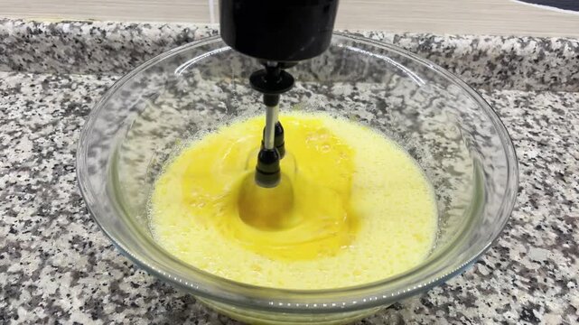 A mixture of egg yolks being whisked with an electric mixer in the kitchen. A close-up shot showing the foamy, runny consistency of the mixture as it is prepared for making a cake or dessert.