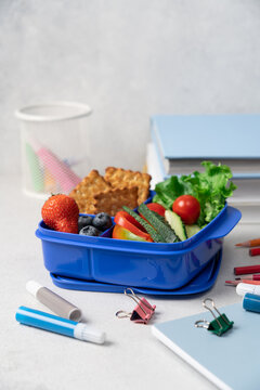 Blue school lunch box with berries crackers and vegetables on light background, side view with stationery, back to school concept