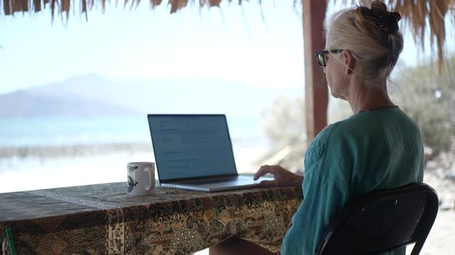 Mature woman sits at a table using a computer under a palapa on a beach in Baja California, Mexico while enjoying the day.