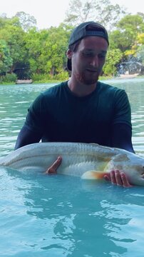 Vertical video. Man Holding Large Catfish In Water Showing Catch Before Release In Lake With Trees And Fountain Background, Recreational Fishing Moment