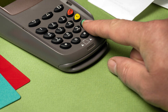 A hand is pressing buttons on a payment terminal placed on a green surface. Nearby are several credit cards. This scene takes place in a retail environment