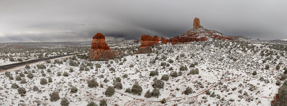 Aerial view of snow-covered red rock buttes and desert landscape under a dramatic overcast sky near a winding road in Page, Arizona, United States.