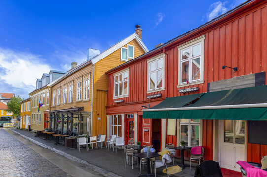 A street view of historic colorful wooden buildings in Trondheim, Norway, featuring red and yellow facades with outdoor cafe seating under a bright blue sky.