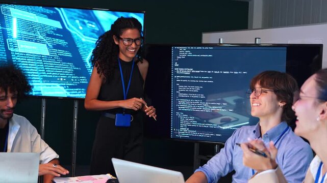 Smiling female programmer presenting computer code and software script on screen to her team. Diverse group of students learning web development and data science in classroom during night workshop