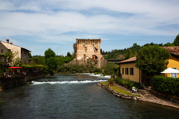 Obraz premium Ancient stone castle ruins and traditional colorful houses along a flowing river in a historic European village under a bright blue sky with wispy clouds captured with a wide angle lens in natural day