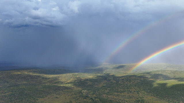 Aerial view of a double rainbow stretching across a vast green landscape under dark storm clouds in Waterberg District Municipality, Limpopo, South Africa.