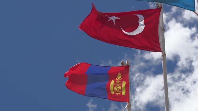 Turkish Mongolian flags waving on white iron pole, national emblems in partly cloudy sky. Turkey Mongolia state pennants fluttering atop mast, bilateral friendship beneath blue clouds.