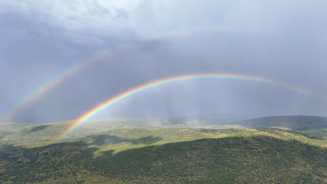 Aerial view of a double rainbow over rolling green hills and dense vegetation under a dramatic cloudy sky in Waterberg District Municipality, Limpopo, South Africa.