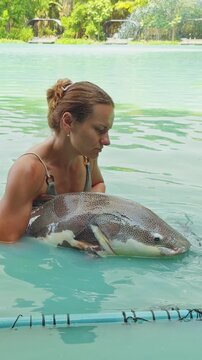 Vertical video. Woman Holding Large Redtail Catfish In Shallow Lake Water Inside Landing Frame With Focused Expression, Careful Fish Handling And Catch And Release Preparation