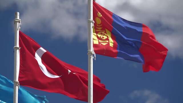 Turkish Mongolian flags waving on white iron pole, national emblems in partly cloudy sky. Turkey Mongolia state pennants fluttering atop mast, bilateral friendship beneath blue clouds.