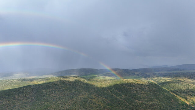 Aerial view of a vibrant double rainbow arching over the rolling green hills and rugged terrain under a dramatic stormy sky in the Waterberg District Municipality, Limpopo, South Africa.