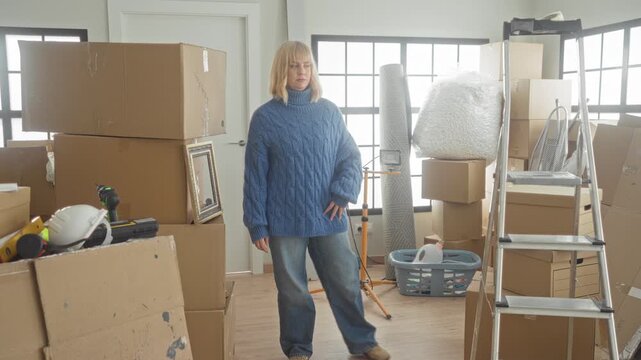 Woman standing with hands on hips among packed cardboard boxes, ladder, bubblewrap and tools in a sunlit building room; moving transition stress.
