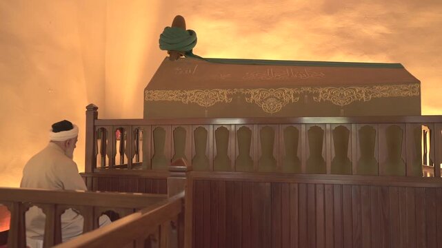 Muslim elder man prays at mausoleum shrine in masjid, Islamic worship in quiet interior light. Aged devotee performs salat near tomb chamber within mosque, traditional cleric wearing robes and turban.