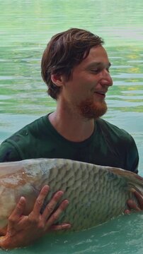 Vertical video. Large Carp Thrashing In Man Arms Causing Strong Splash And Knocking Cap Off Head, Dynamic Fishing Struggle And Action Moment