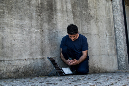 A man in blue workwear squats against a concrete wall, using his mobile phone to configure or monitor a portable solar panel and LED light system.