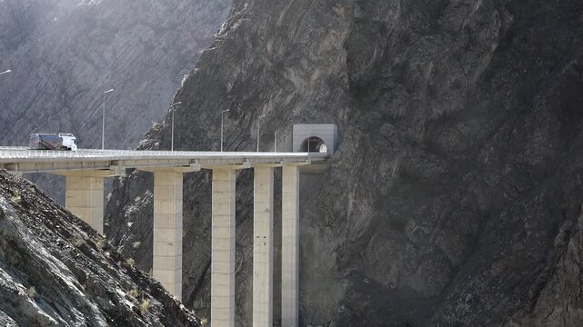 High concrete viaduct bridge and tunnel on mountain highway in Himalayas rocky valley. Dangerous elevated road span with pillars near cliff gorge, linking route to bore entrance.