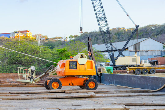 Orange telescopic boom lift on a wooden pier with a port crane in the background in Willemstad Curacao