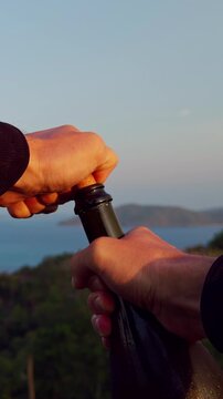 Vertical video. Hands Opening Champagne Bottle With Cork Against Scenic Sea And Hills Background At Sunset, Celebration And Luxury Leisure Moment