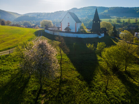 Aerial view of the Gothic Church of Saint Martin with its wooden bell tower and white defensive wall amidst green hills and blooming trees Cerin, Banskobystricky kraj, Slovakia.