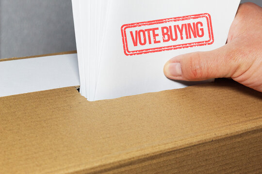 A hand inserting several ballots stamped Vote Buying into a cardboard ballot box, a direct depiction of electoral fraud in the electoral process