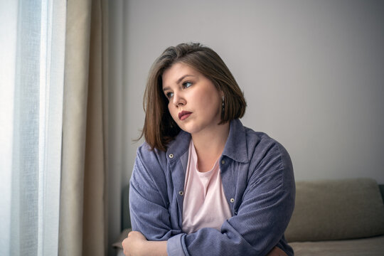 Thoughtful plus size woman looking through window, sad home portrait, self reflection, mental health, emotional wellness, inclusive lifestyle.