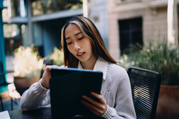 Fototapeta premium Asian woman sits at outdoor café, gently smiling while using digital tablet. Scene blends relaxation with seamless tech interaction and modern solo work culture.