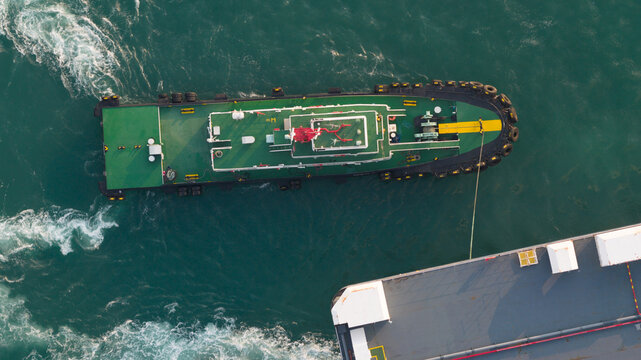 Aerial view of a green tugboat maneuvering alongside a large vessel in the turquoise waters of the industrial port in Laem Chabang, Chon Buri, Thailand.