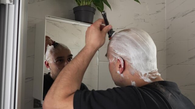 Bald man shaving his beard and head at home in front of a mirror