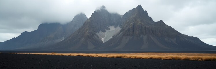 Naklejka premium Jagged grey mountains with snow patches rise above dark volcanic sand. Golden grass borders the desolate landscape under a cloudy sky. This wild nature scene offers immense scale and solitude.