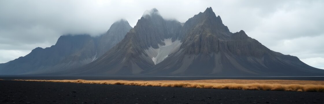 Jagged grey mountains with snow patches rise above dark volcanic sand. Golden grass borders the desolate landscape under a cloudy sky. This wild nature scene offers immense scale and solitude.